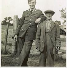 A tall, youthful BG Sgt. in full uniform leans with his arm resting on the shoulder of the little, old farmer. They both smile.